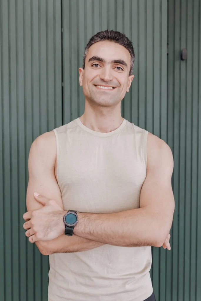 Smiling man wearing a beige sleeveless shirt and a black watch, standing in front of a green corrugated metal wall.