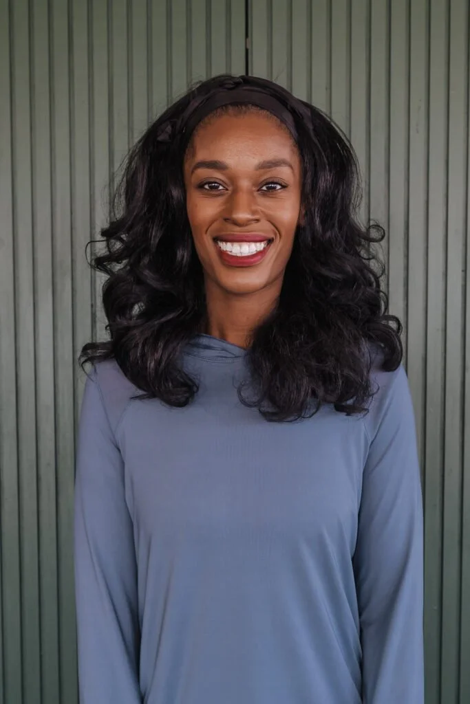 Smiling woman with long, wavy black hair wearing a dark headband and a gray long-sleeve top, standing in front of a vertical gray-green fence.