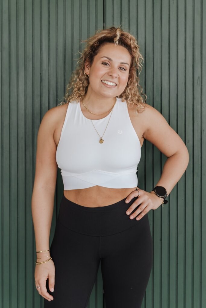 Woman smiling, wearing a white sleeveless crop top, black high-waisted pants, gold jewelry, and a black smartwatch, standing against a green paneled wall.