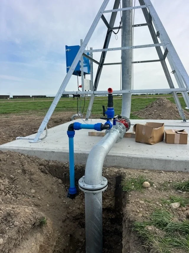 A water pipe system installed at the base of a wind turbine tower on a concrete foundation, with blue valves and fittings, surrounded by soil and grass in an open field.
