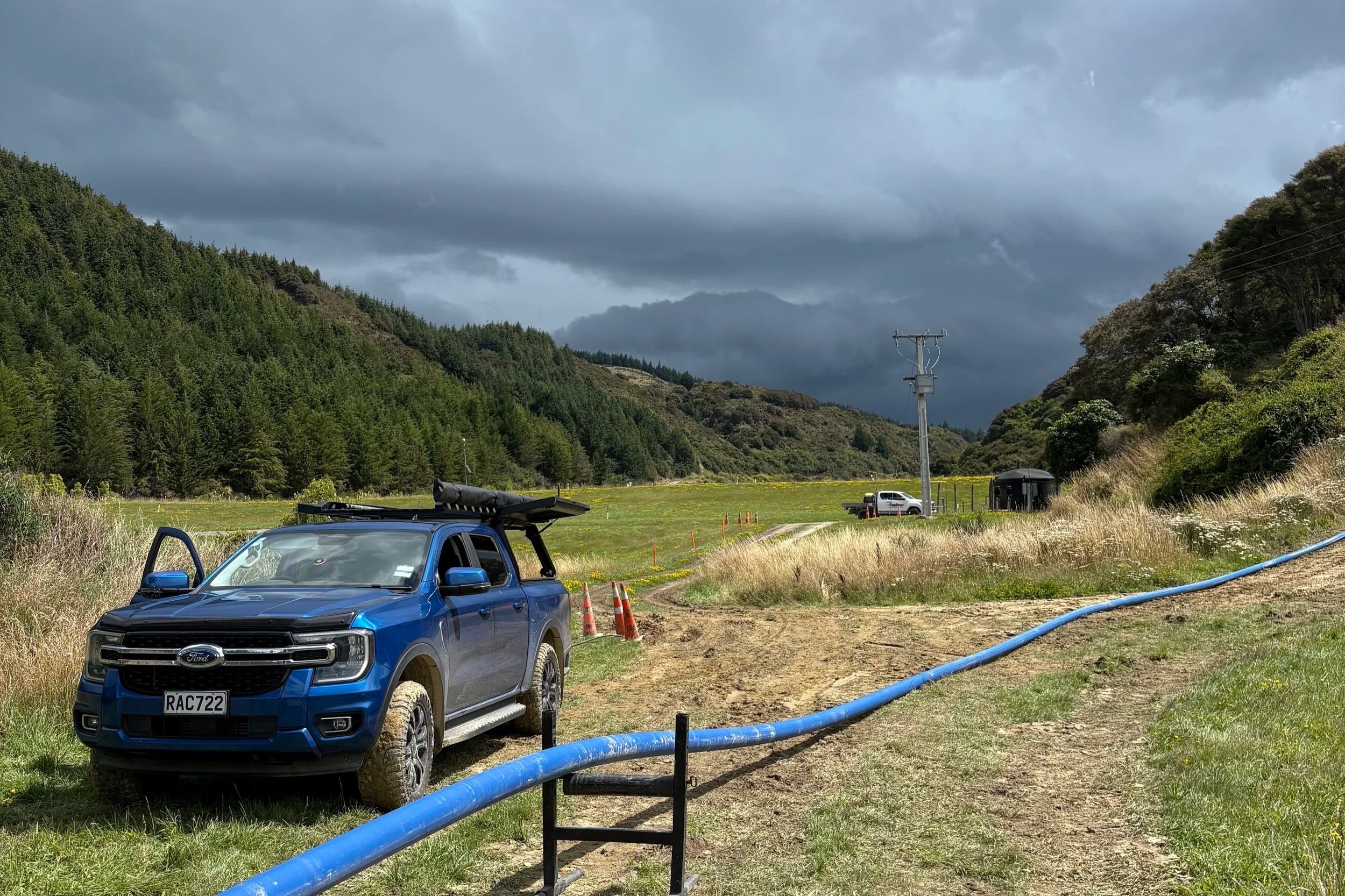 A scenic outdoor landscape with rolling hills covered in trees, a cloudy sky, utility poles, cars, and construction cones on a dirt road with a blue hose on the ground.