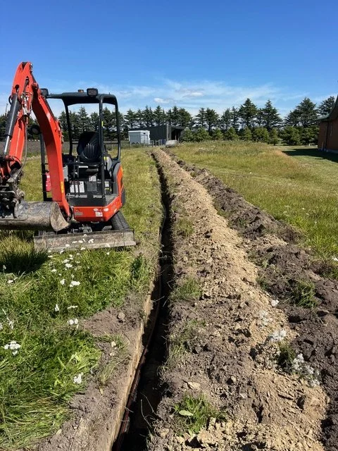 A mini excavator digging a trench in a grassy yard on a sunny day with a clear blue sky.
