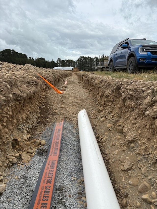 A construction site showing a trench with a large white pipe and a warning marker on an orange cable. A blue pickup truck is parked nearby under a cloudy sky.