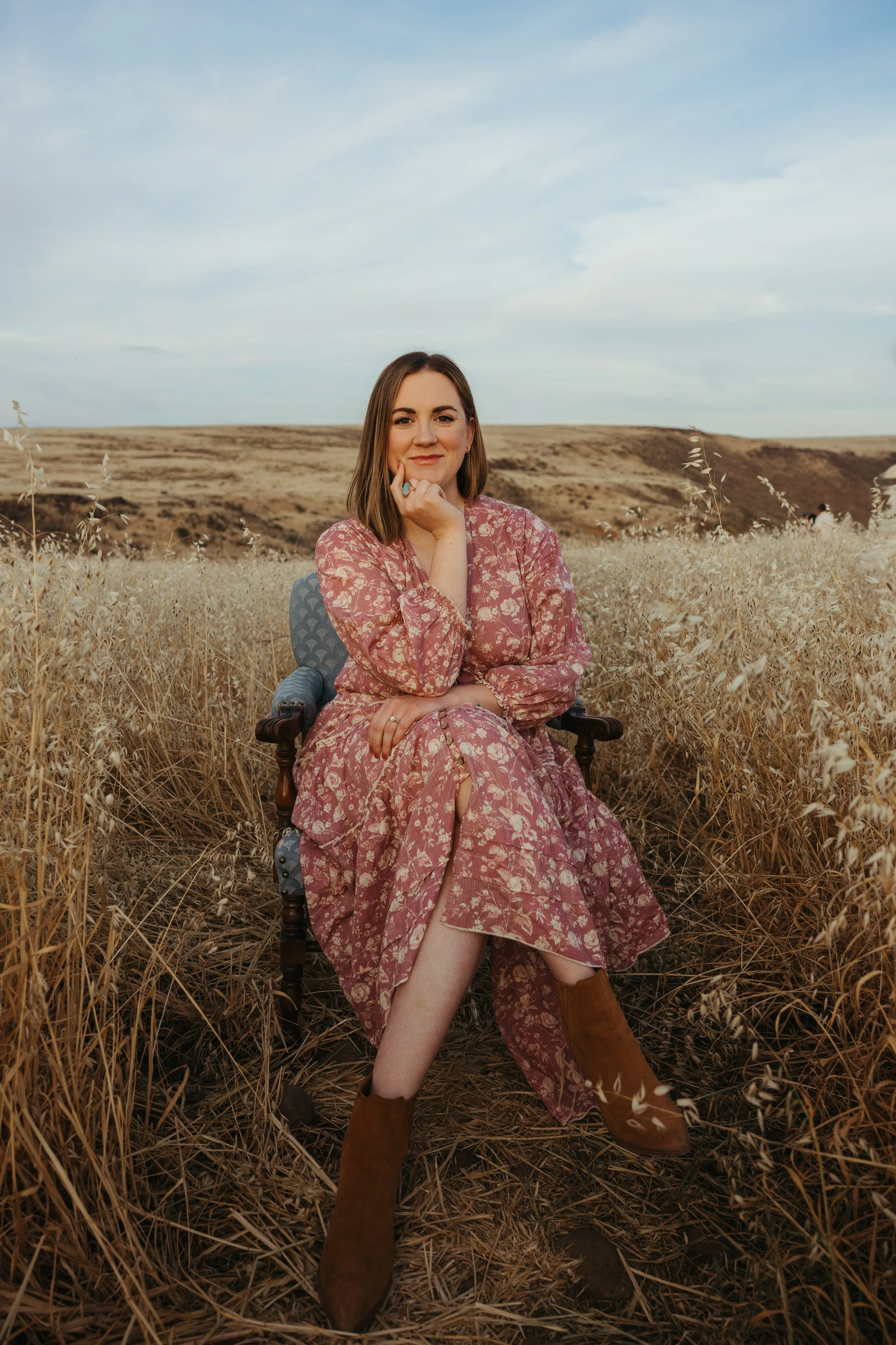 A female perinatal, women's and trauma therapist with brown hair and light skin, wearing a pink floral dress in a chair in a field of tall, dried grass. She is smiling, her hand resting on her chin, and the background features rolling hills and sky.