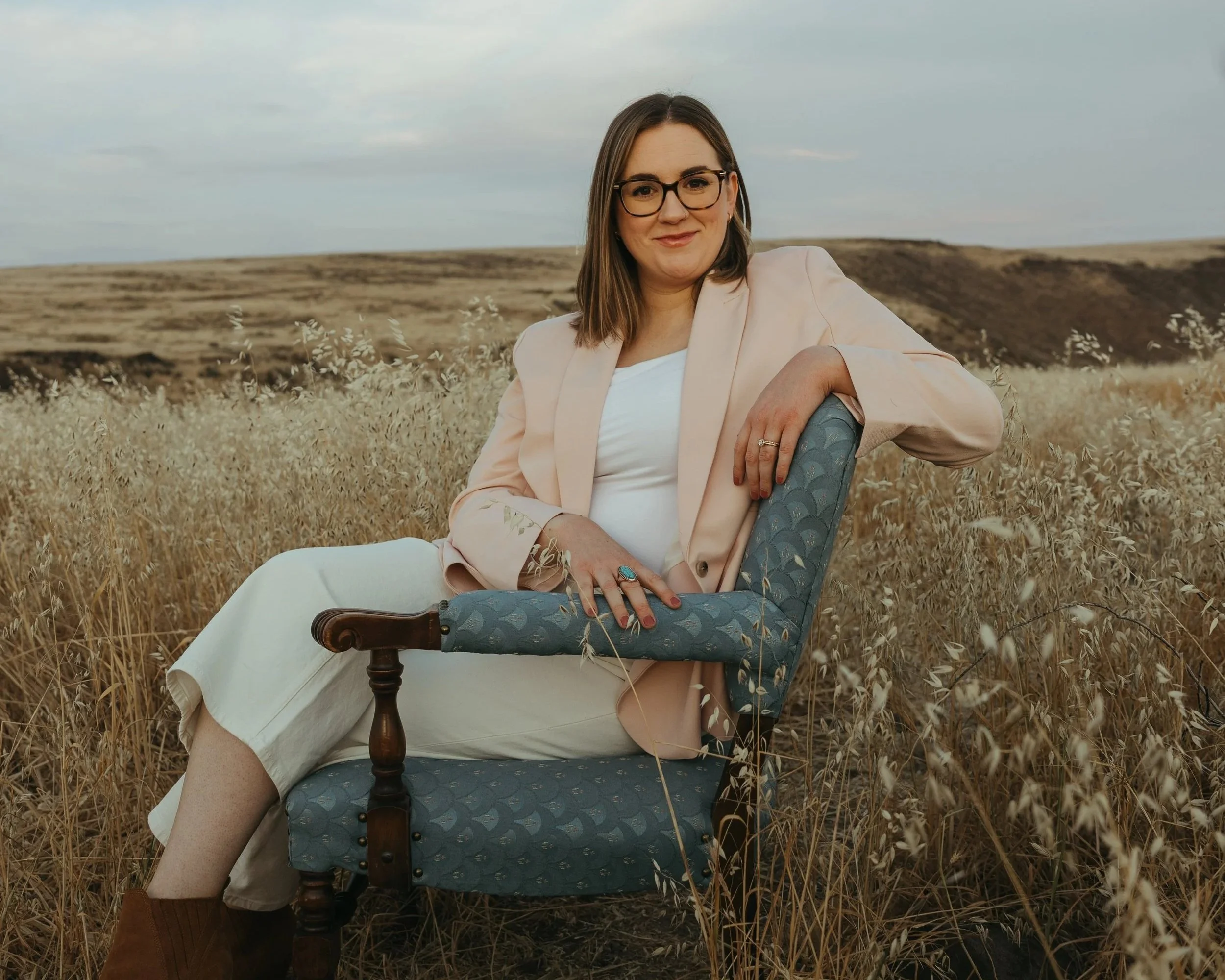 A female perinatal, women's and trauma therapist with glasses, brown hair, and a light pink blazer sitting on a vintage chair in a field of tall grass, with rolling hills and a cloudy sky in the background.