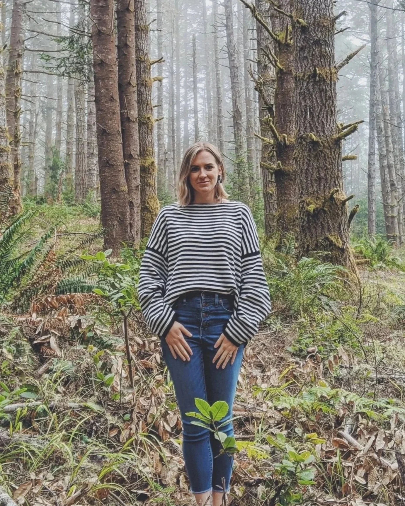 A woman standing among trees and green foliage in a forest.