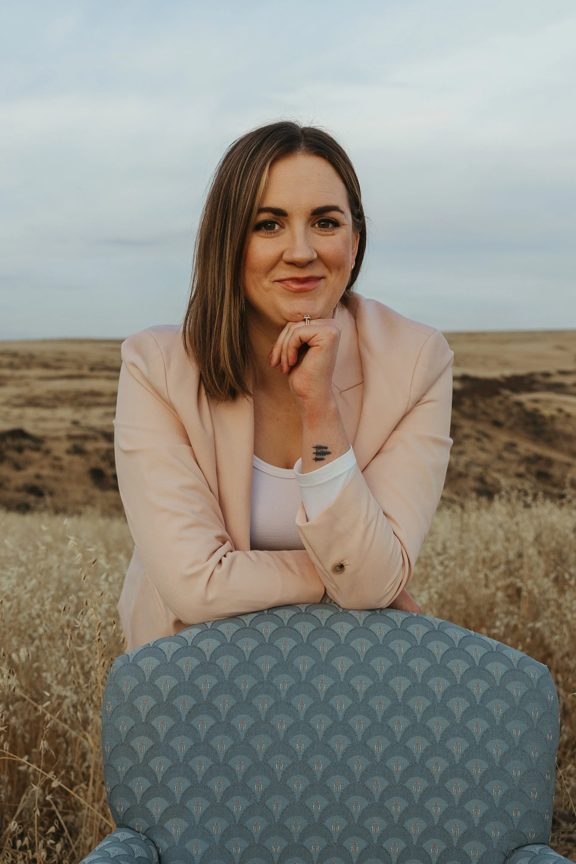 A female perinatal, women's and trauma therapist with shoulder-length brown hair, wearing a light pink blazer and white top, is leaning on a pastel blue chair in a dry, open field during daylight, with a cloudy sky in the background.