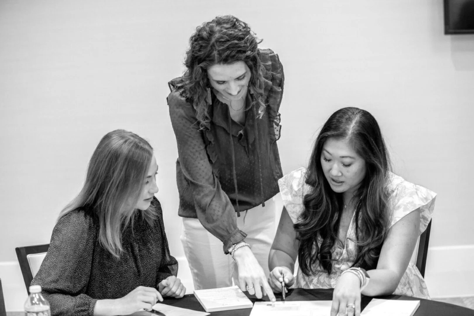 Three women are gathered around a table, reviewing documents and a tablet. One woman is standing and pointing at something on the table, while the other two women are seated, looking at the documents and tablet.
