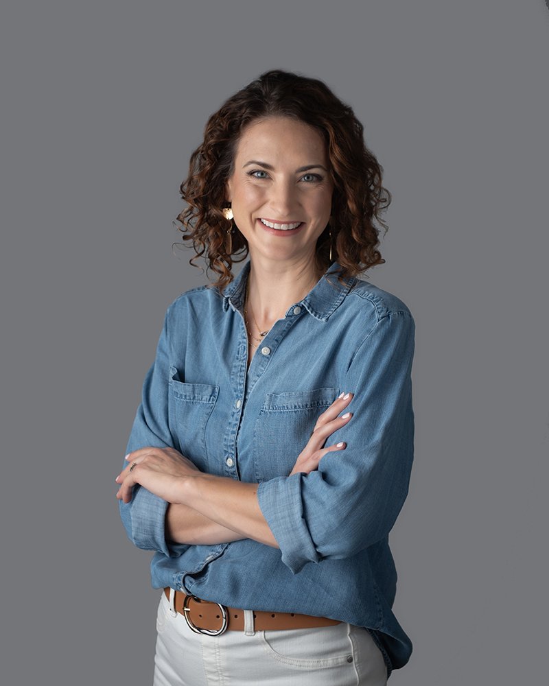 A smiling woman with curly brunette hair, wearing a denim button-up shirt and white pants, standing against a gray background.