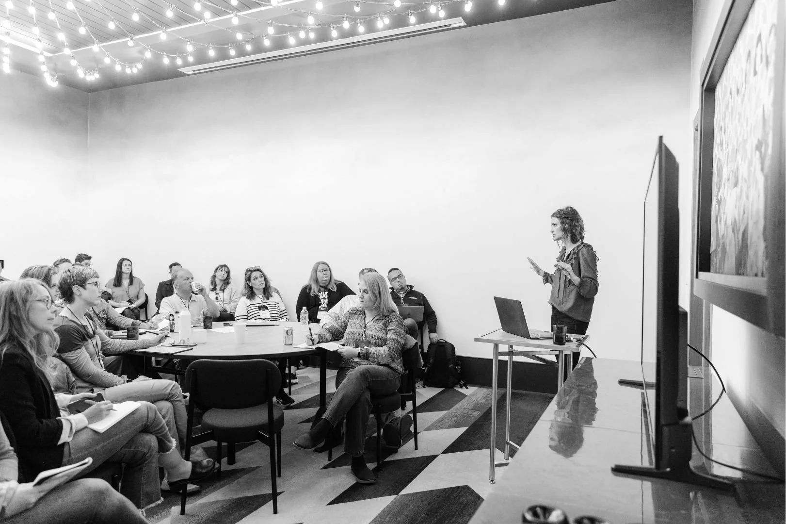 A woman giving a presentation to an audience in a conference room with string lights on the ceiling, some attendees taking notes, and a flat-screen TV on the wall.