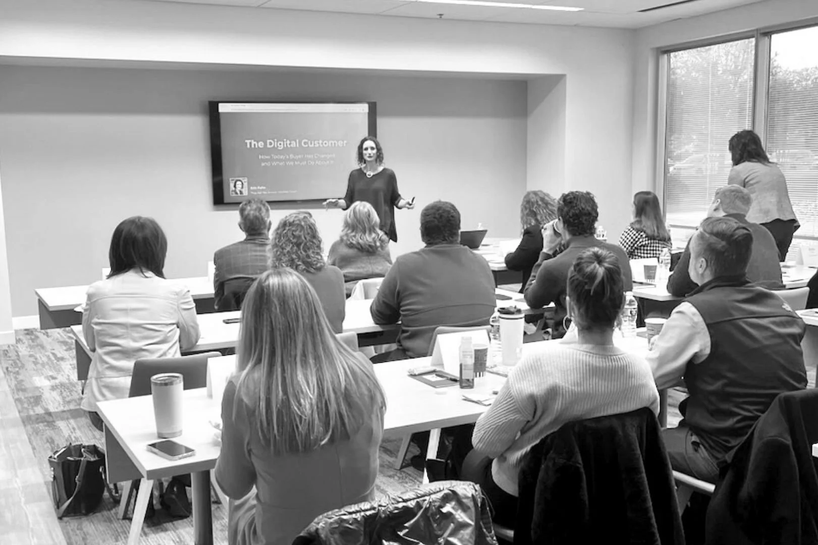 A woman presenting to a group of adults in a conference room with a screen that reads "The Digital Customer." The audience is seated at tables with notebooks and drinks, attentively listening.