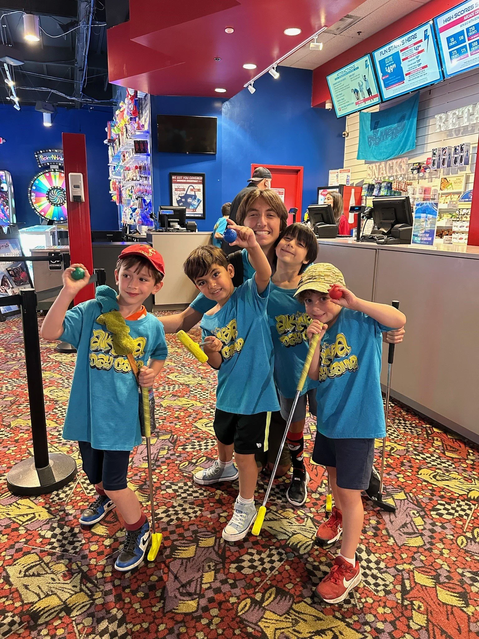 A group of five children and one adult boy smiling and holding colorful balls and mini golf clubs inside an arcade or entertainment center with a red and blue color scheme, a game wheel, and counter screens visible in the background.