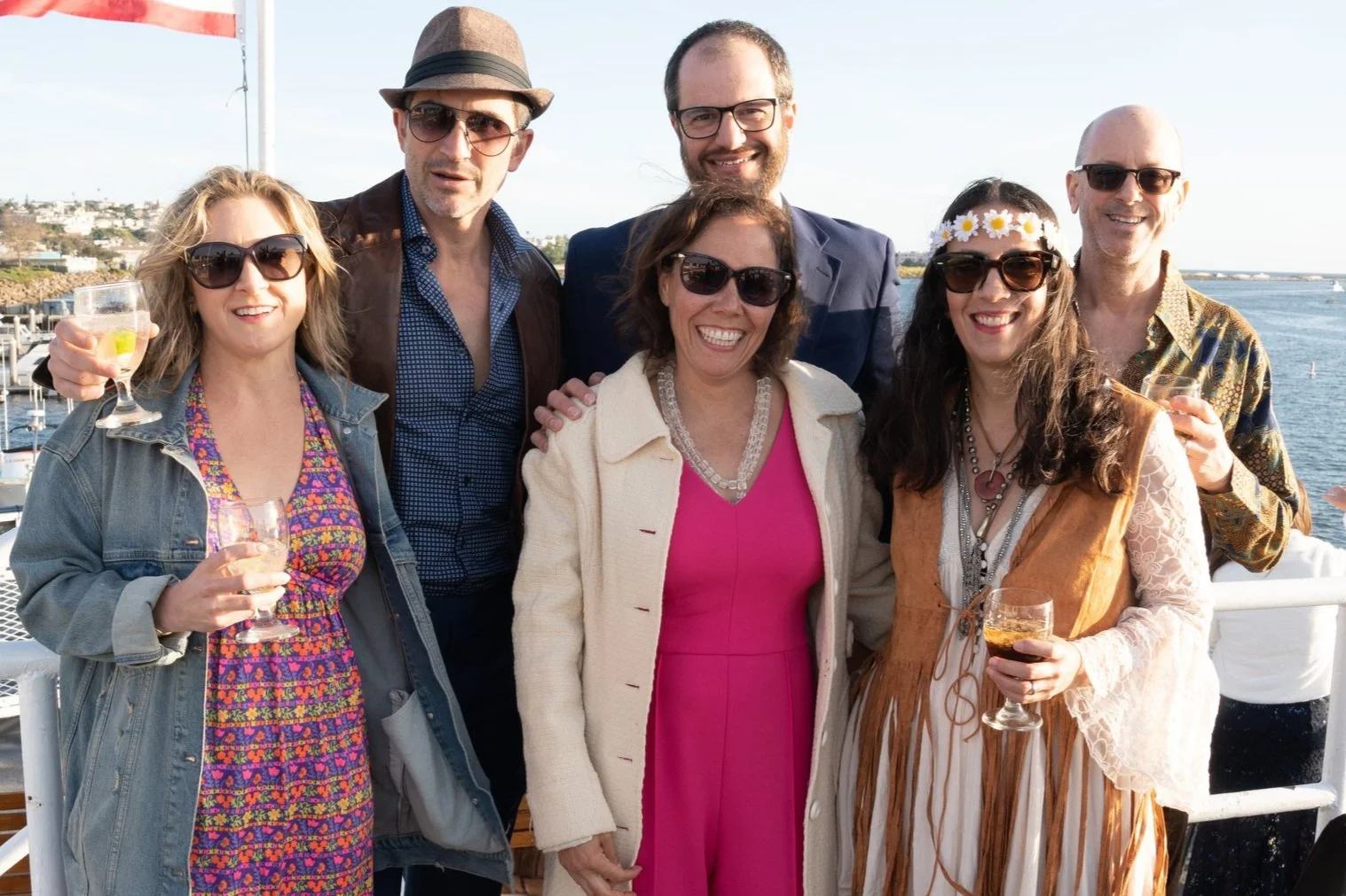 Group of seven adults smiling on a boat deck with water and a shoreline in the background, holding drinks and dressed casually for warm weather.