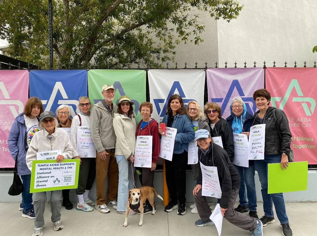 Group of people participating in a mental health walk event, holding signs supporting mental health awareness, standing outdoors in front of colorful banners with the logo of a mental health organization.