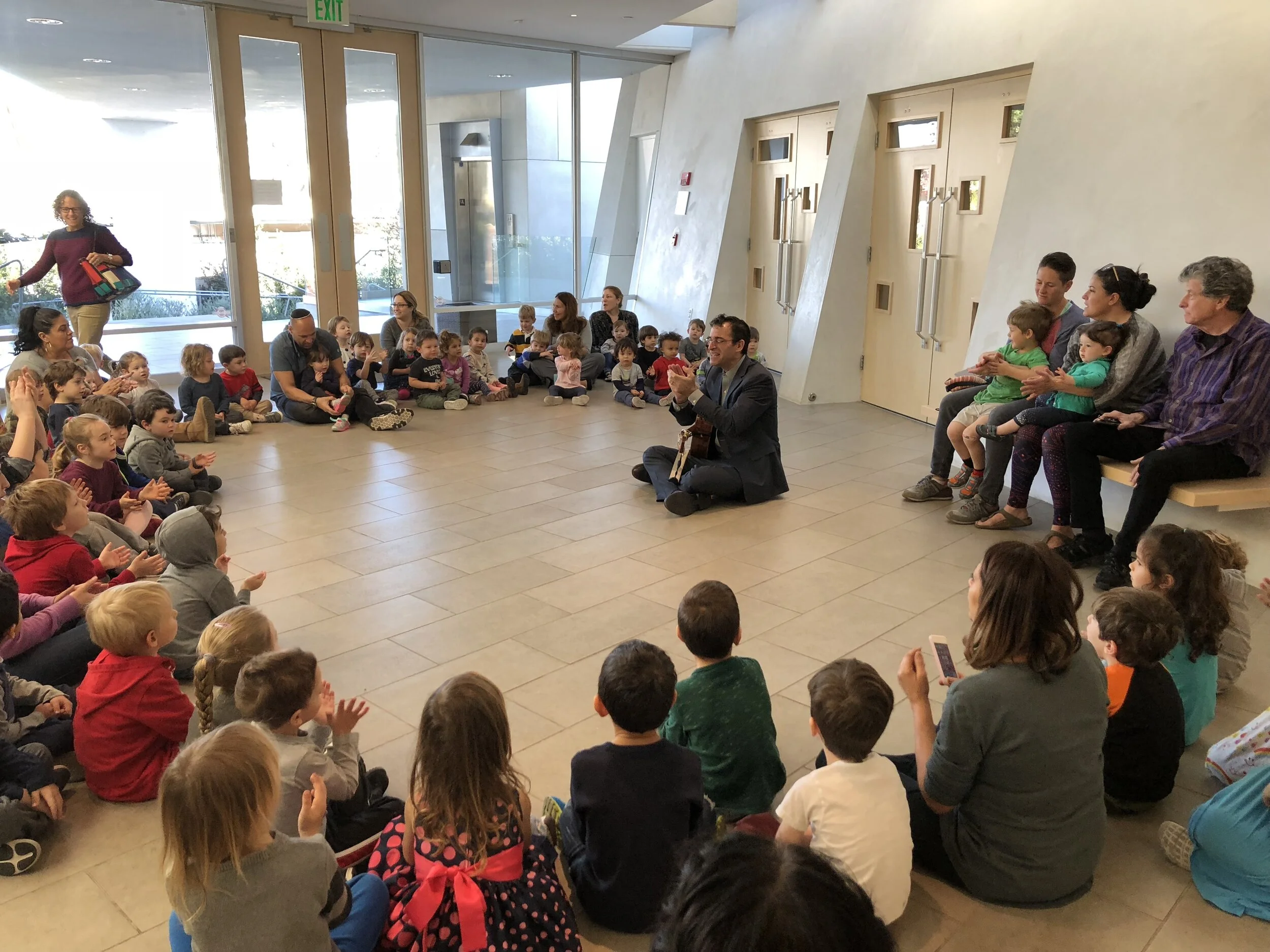 A man playing guitar and singing seated on the floor in front of a large group of children and some adults sitting on the floor and benches in a spacious, modern indoor setting, with glass doors and windows in the background.