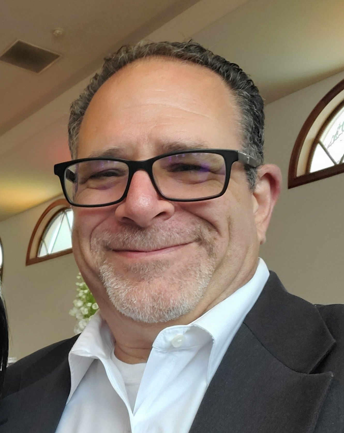 Smiling man wearing glasses, white dress shirt, and black suit, indoors with arched windows in the background.