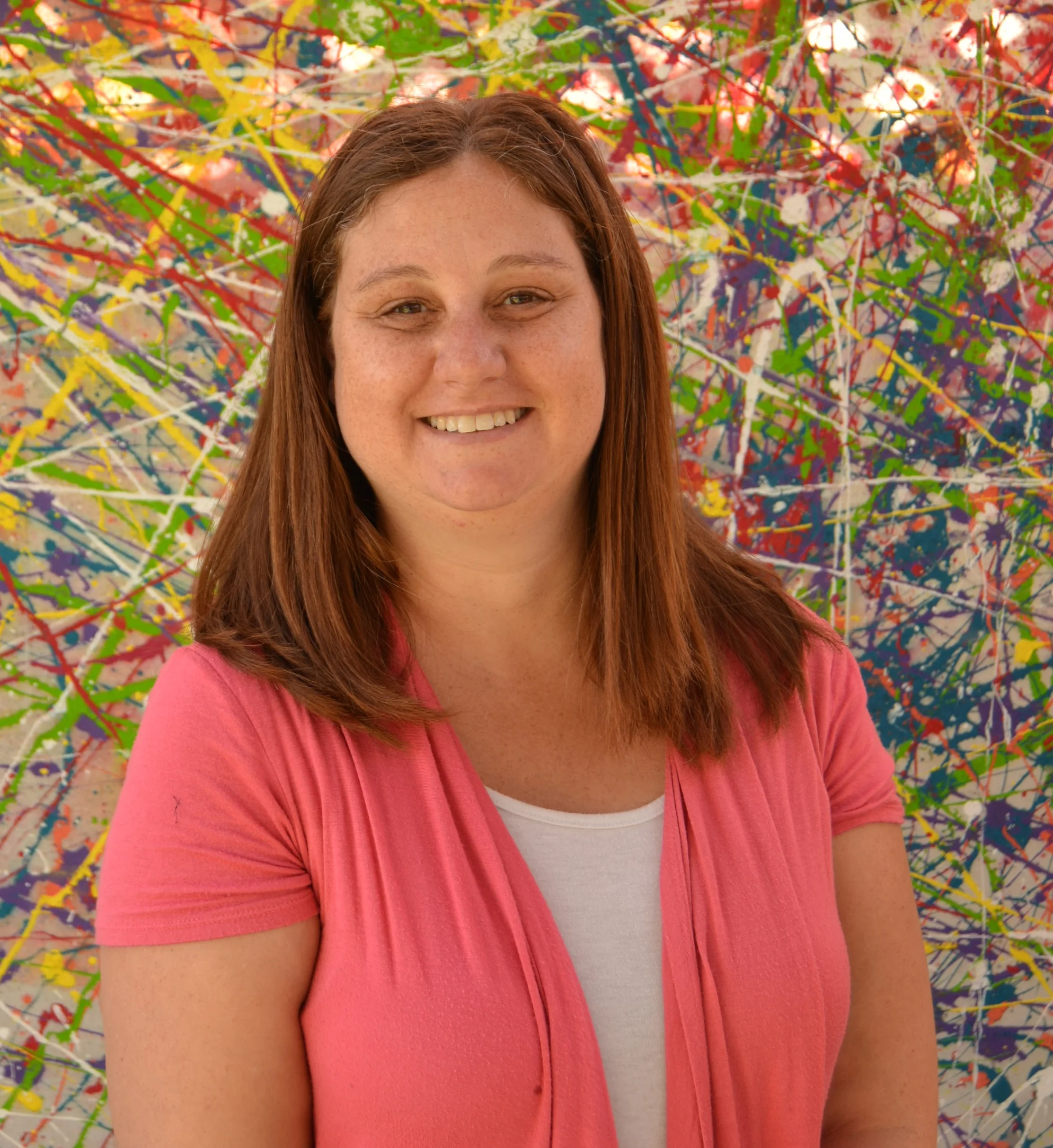 A smiling woman with shoulder-length reddish-brown hair wearing a pink shirt, standing in front of a colorful abstract background with splattered paint in various colors.