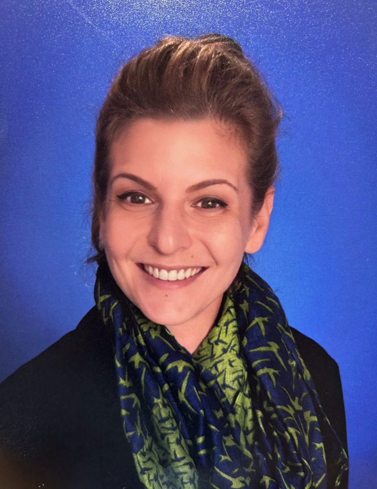 A woman smiling with short brown hair, wearing a black top and a blue and green patterned scarf, against a blue background.