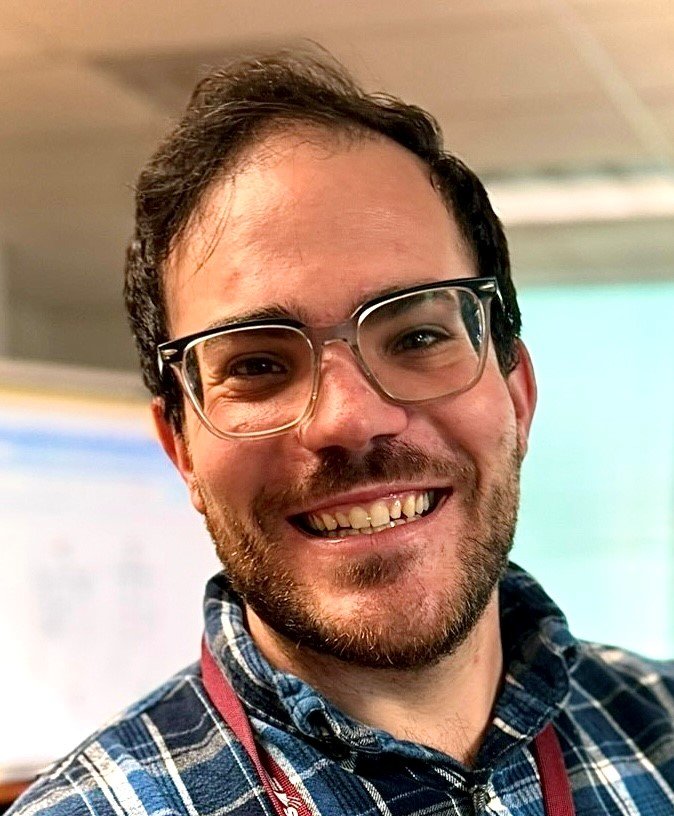A smiling young man with glasses and a beard, wearing a plaid shirt indoors.