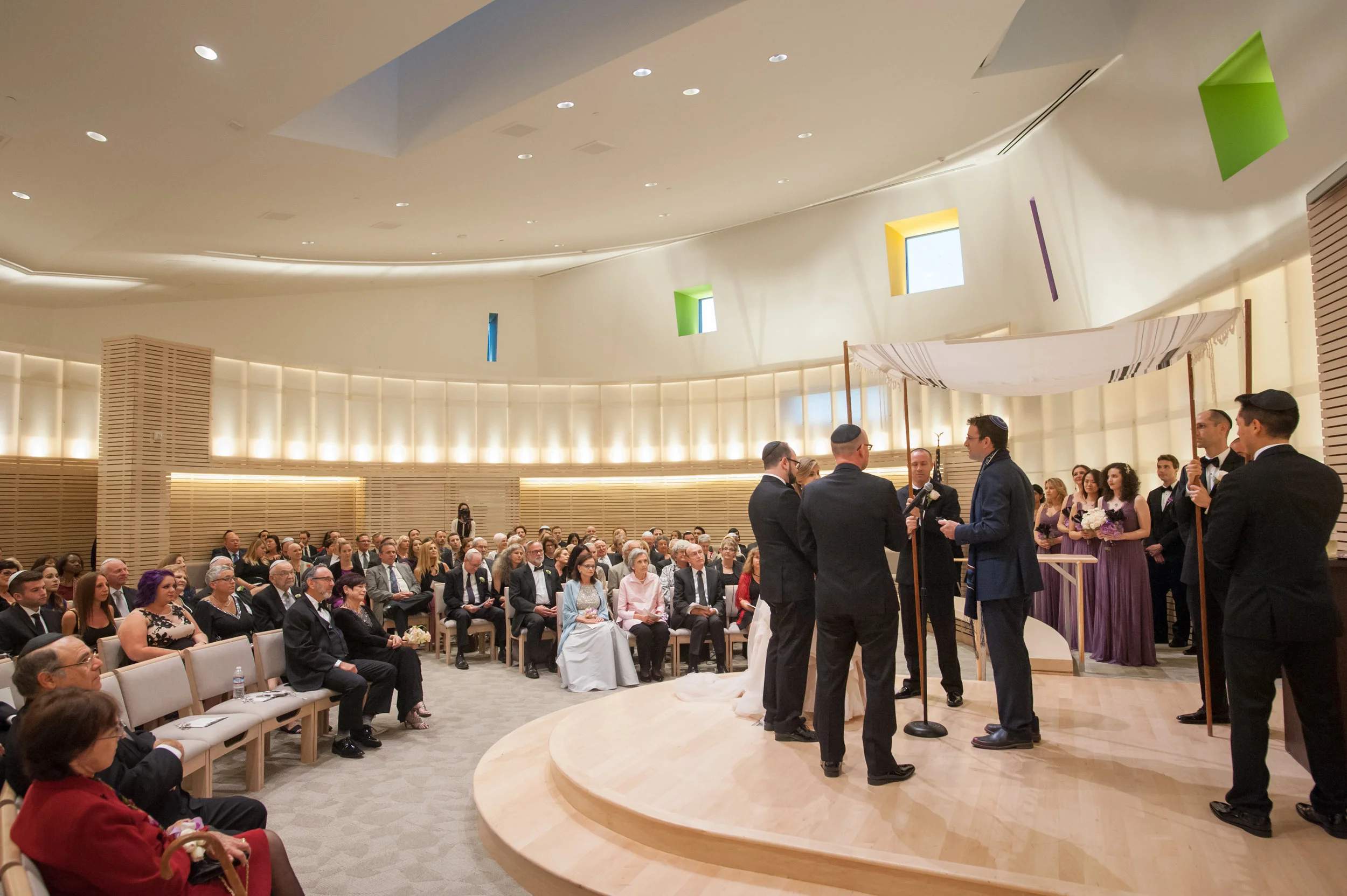 A wedding ceremony in a modern indoor venue with clergy officiating, surrounded by the bride and groom, wedding party, and guests seated in rows. The venue has rounded walls, colorful window accents, and overhead lighting.