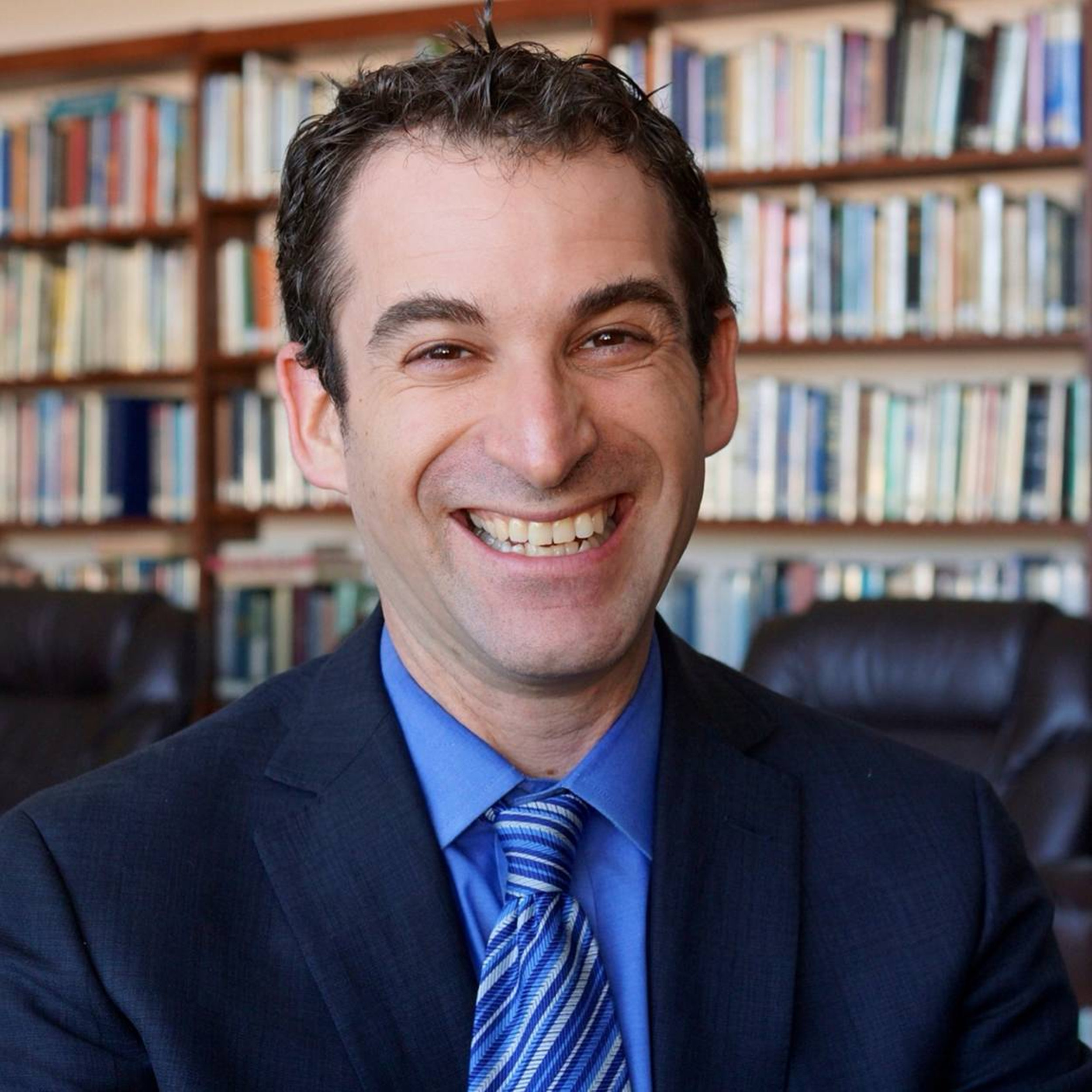 A smiling man in a suit and tie in a library setting, with bookshelves in the background.