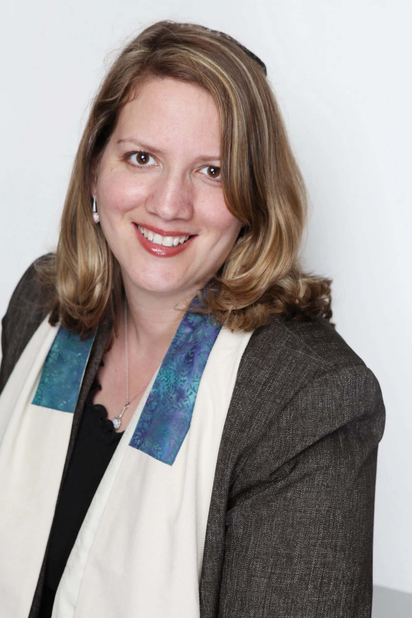 Headshot of a smiling woman with shoulder-length light brown hair, wearing a black top, brown jacket, a white scarf with a colorful pattern, and earrings.