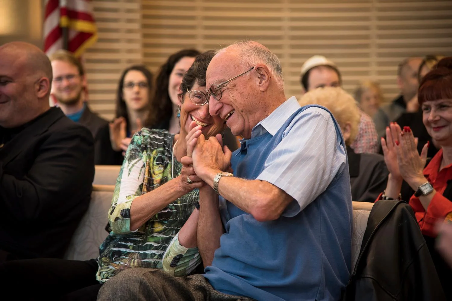 An elderly couple sits in a crowded auditorium, smiling and laughing as they hold hands, surrounded by other applauding attendees.