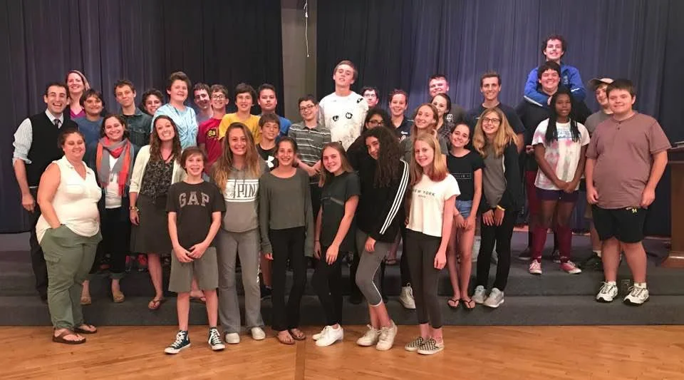 Group of students and adults posing on a stage, smiling for a photo in a school or auditorium setting.