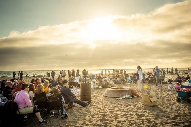 Crowd of people gathered on a beach at sunset, sitting and standing around, with some near a fire pit and others along the shoreline.