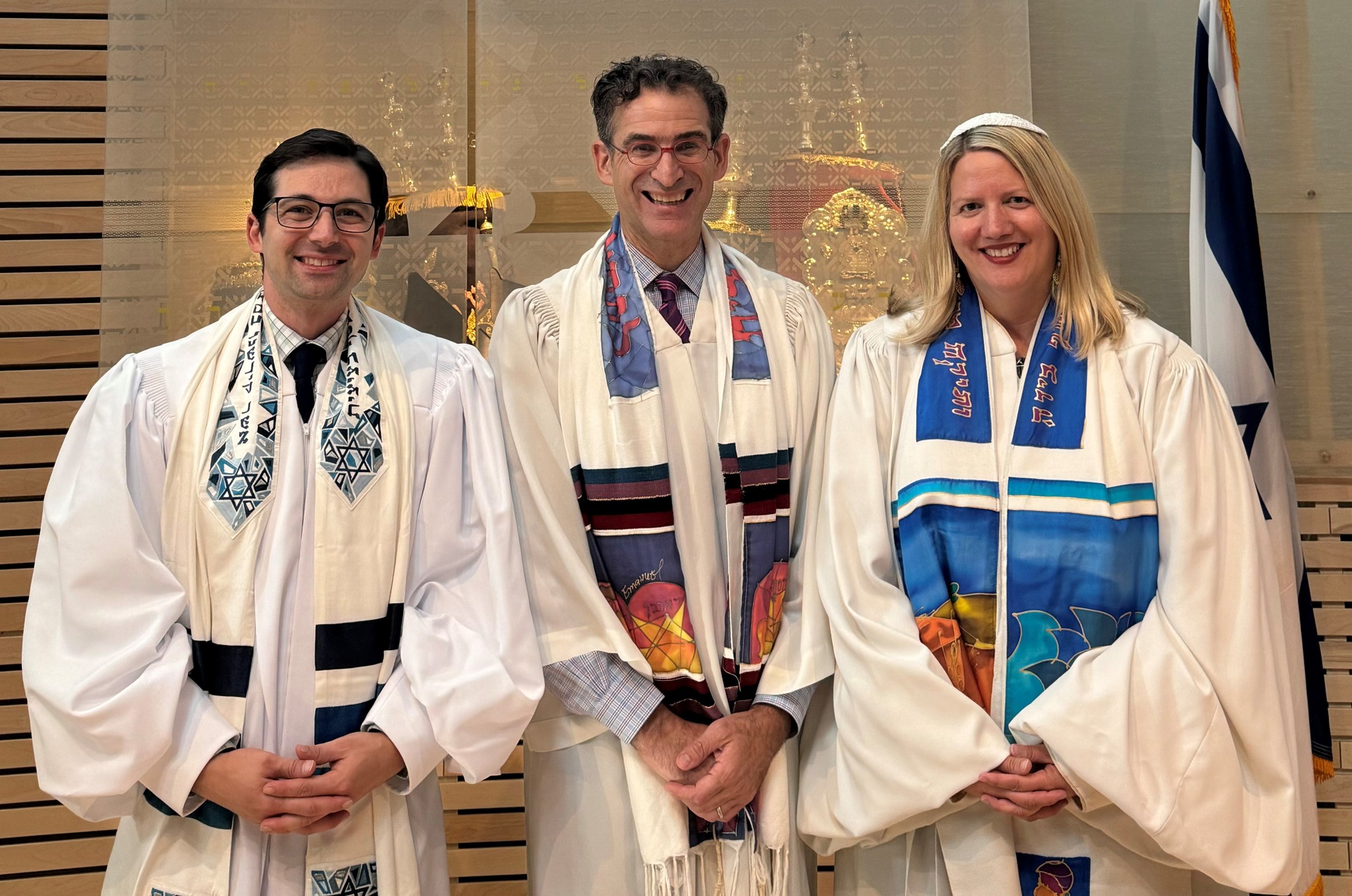 Three individuals dressed in religious garments, standing indoors for a ceremonial event, with Jewish symbols and flags in the background.