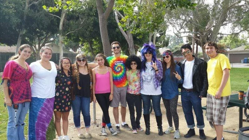 Group of ten young people standing outdoors in a park with trees and a bench in the background, dressed in colorful and quirky outfits, some with wigs and tie-dye shirts, smiling and posing for a photo.