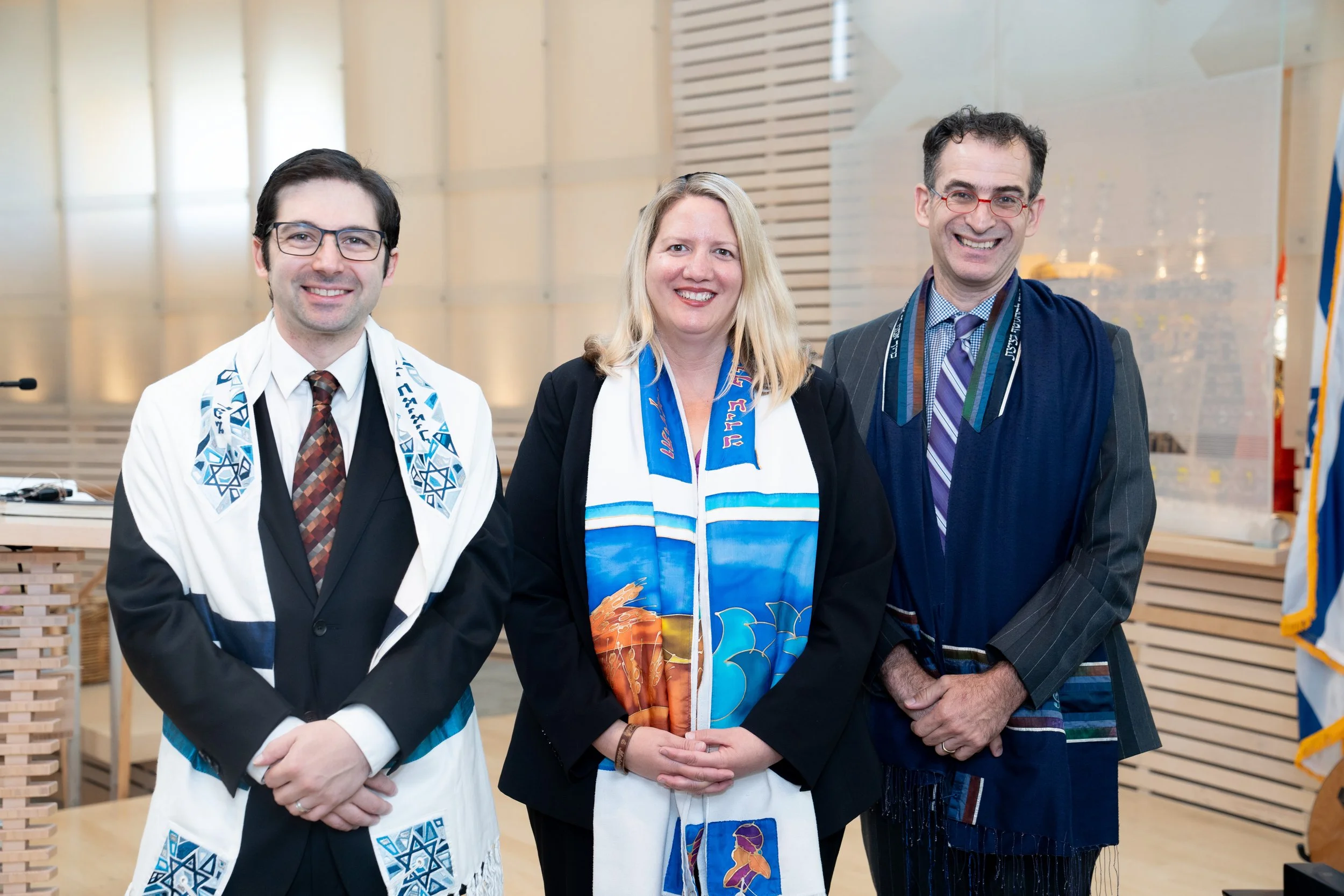 Three individuals standing together indoors, smiling, wearing ceremonial scarves and garments related to a Jewish event or celebration, with a wooden background and a flag in the background.