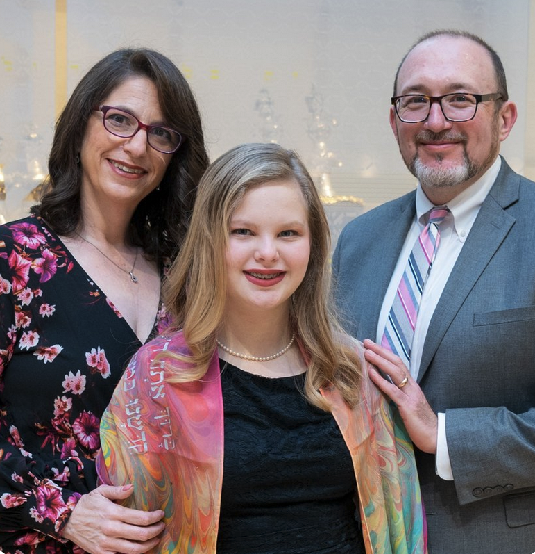 A family of three posing together at an indoor event. The woman on the left wears glasses and a black floral dress, the girl in the middle has long blonde hair, wears a pearl necklace and a colorful shawl over a black dress, and the man on the right has glasses, a beard, a suit, and a striped tie.