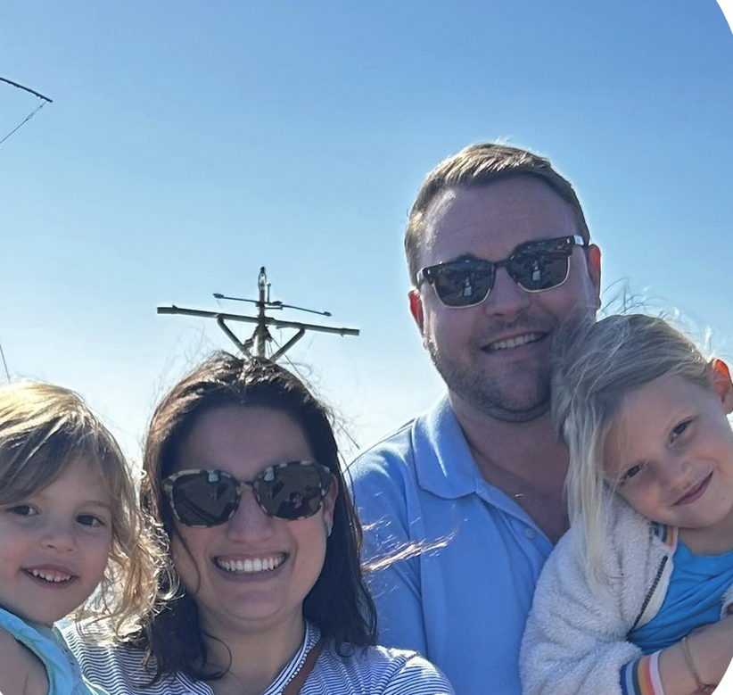 A family of four smiling outdoors on a sunny day with clear blue sky, with a power line in the background.