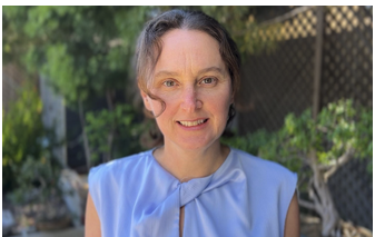 Portrait of a woman with short brown hair and a light blue sleeveless top, smiling outdoors with greenery and a wooden fence in the background.