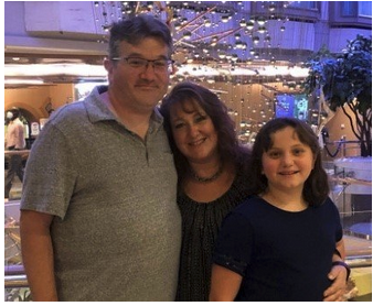 A man, woman, and young girl smiling together at an indoor event, with string lights and a festive atmosphere in the background.