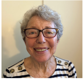 Close-up of an elderly woman with short, curly gray hair, wearing glasses and a striped shirt, smiling at the camera.