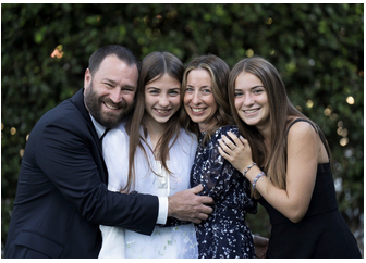 A family of four smiling and embracing outdoors in front of green foliage.