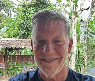 Close-up of a smiling man with gray hair outdoors, background of greenery and a thatched roof structure.