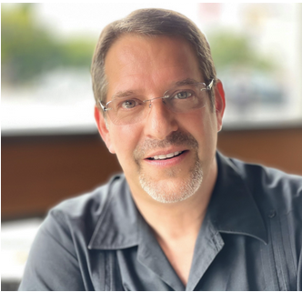 Close-up of a middle-aged man with glasses, smiling, indoors with a blurred window in the background.