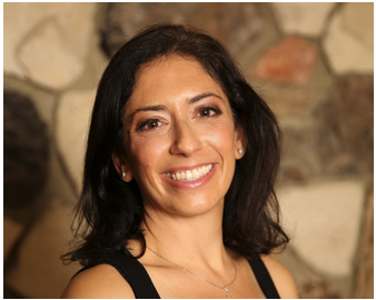 A woman with dark hair smiling in front of a stone wall.
