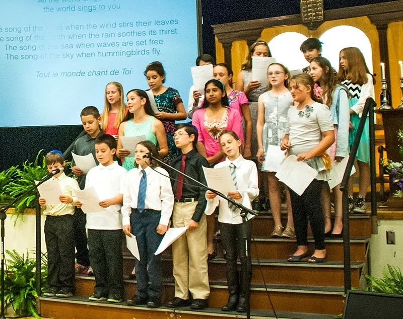 Group of children and teenagers singing on a church or school stage, holding sheet music, with projection screen to the side displaying lyrics.