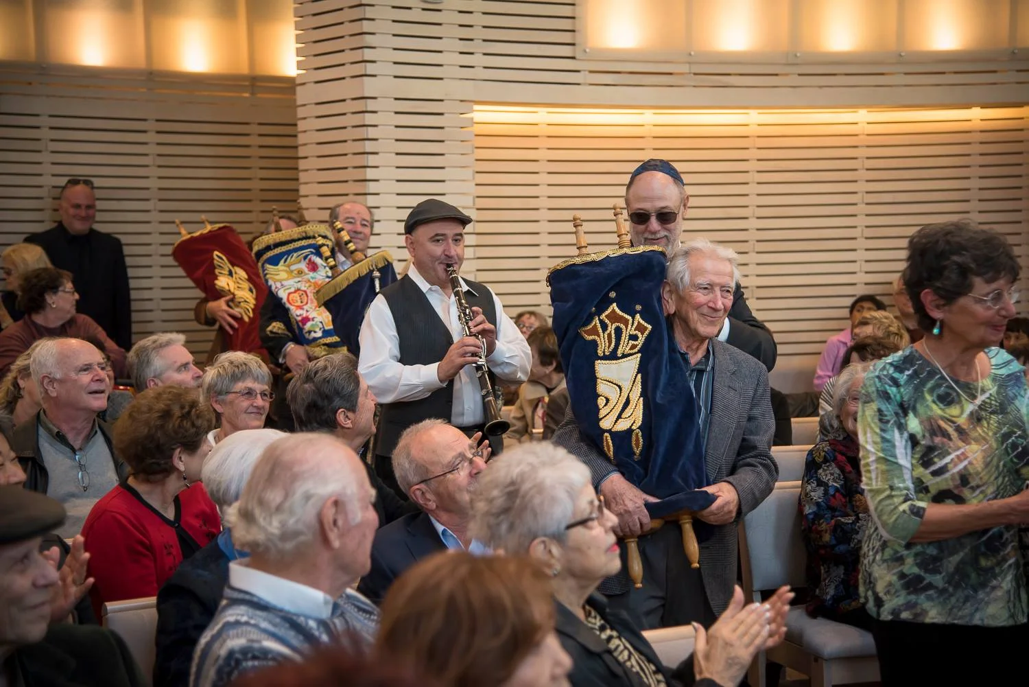 People attending a ceremony or celebration, with some holding flags and banners, one person playing a clarinet, and an audience clapping, in an indoor setting with wooden panel walls.