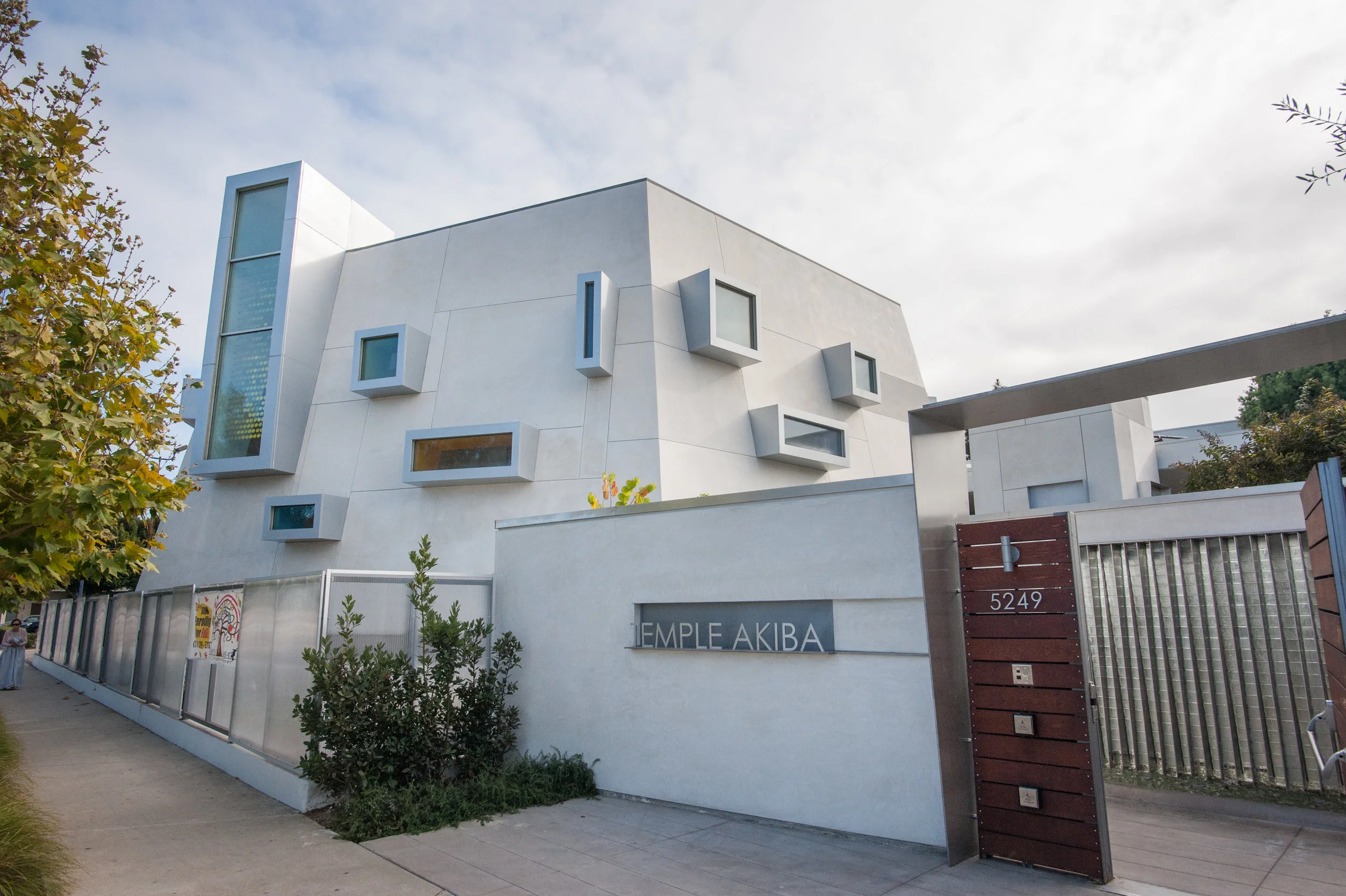 Modern white building with geometric windows and a sign that reads 'TEMPLE AKIBA,' located at 5249, with a sidewalk and trees nearby.