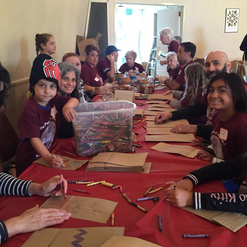 Children and adults gathered around a long table, decorating paper bags and writing messages during a community event.