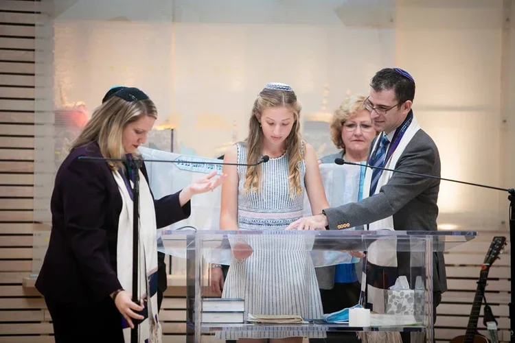 Four people, including a girl in a light blue dress, are gathered around a clear podium with religious books, in a synagogue. They are wearing kippahs and are engaged in a ceremony or prayer.
