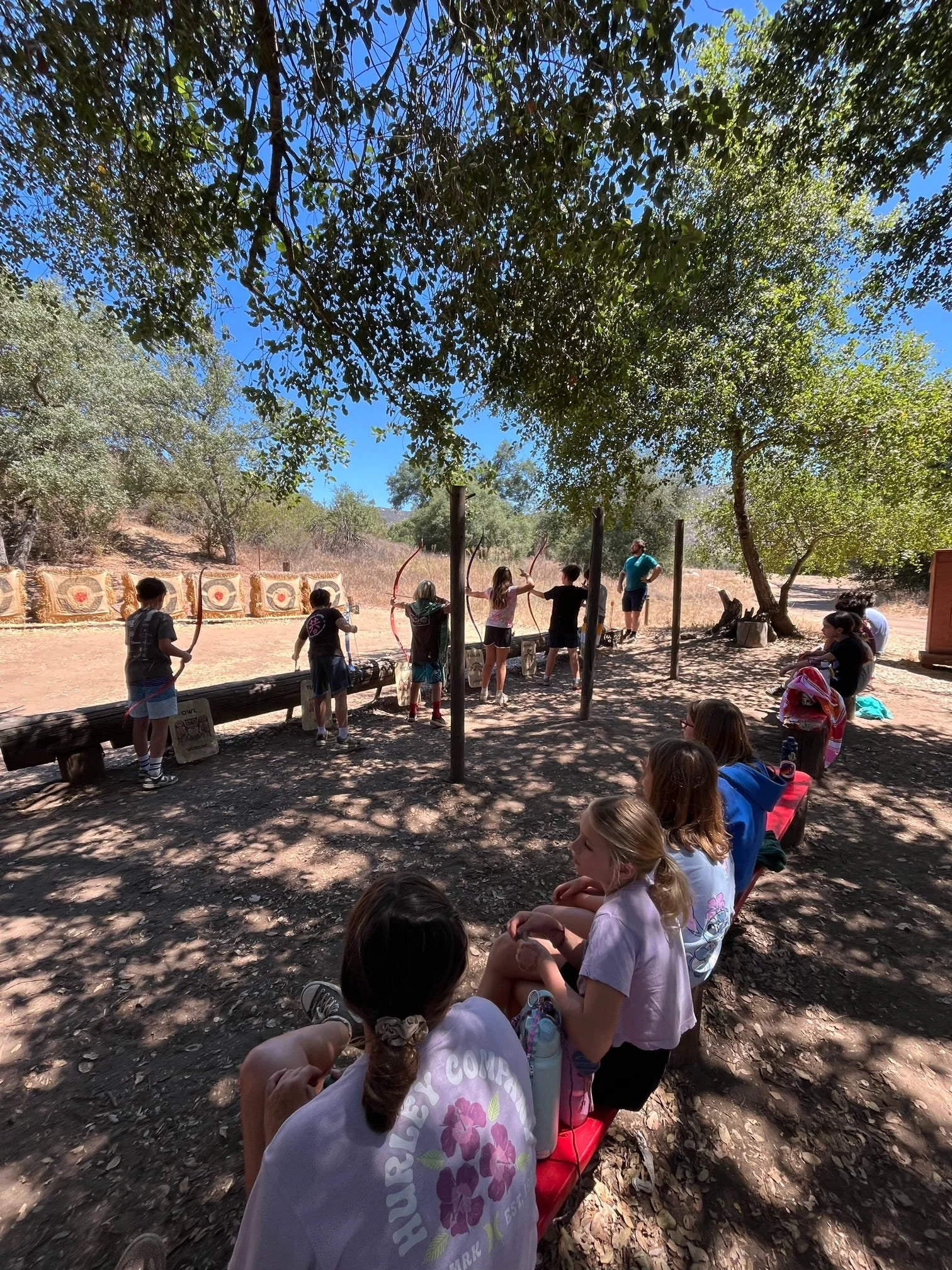 Children participating in outdoor archery activity while seated and standing under trees, with targets and an instructor in the background.