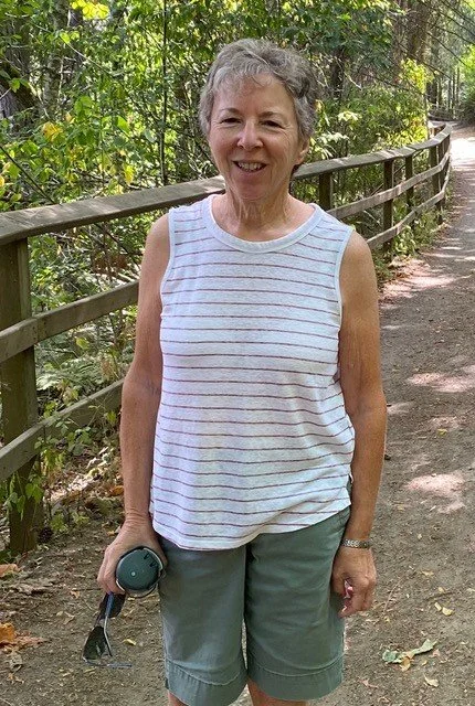 Close-up of an elderly woman with short, curly gray hair, wearing glasses and a striped shirt, smiling at the camera.