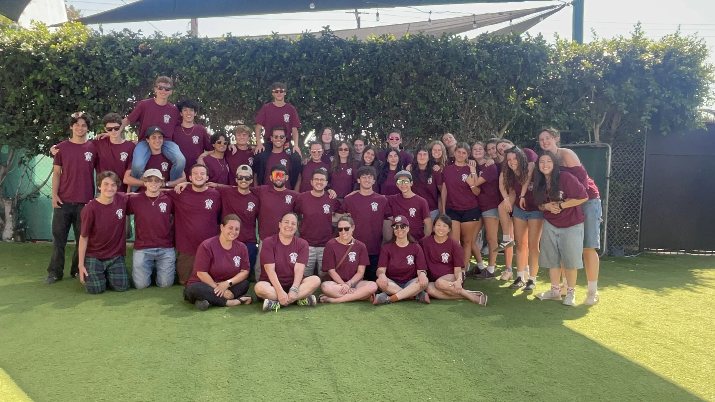 A large group of young people posing outdoors on a grassy field, many wearing matching maroon T-shirts with a logo, smiling at the camera.