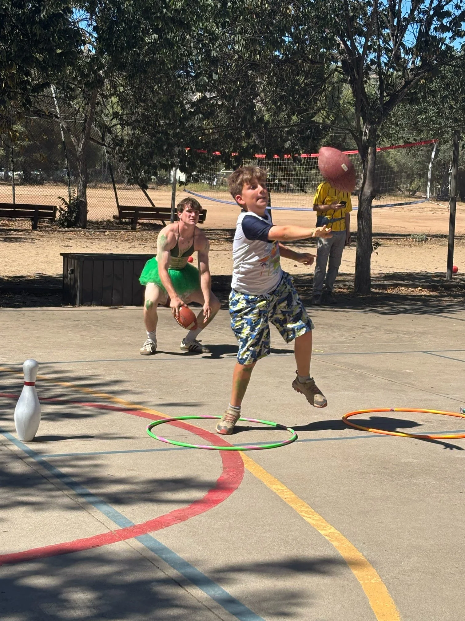 Children playing mini-game with hoops and bowling pin at outdoor recreational area with trees and benches in background
